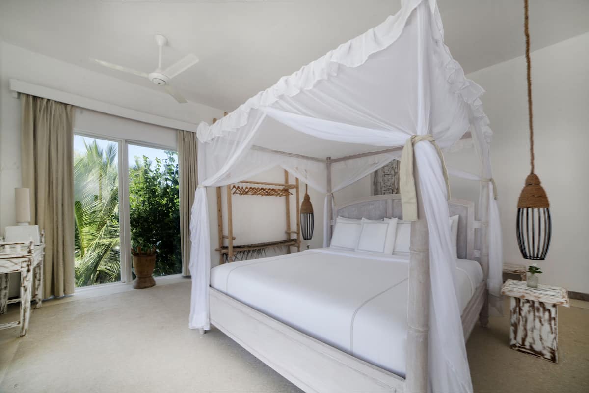A four-poster canopy bed with a white canopy, a ceiling fan, and a view of palm trees through large windows.