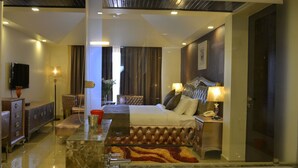 A modern hotel room with a large bed, a sitting area with a sofa and armchairs, a flat-screen TV, a desk with a lamp, and a bathroom with a bathtub. - Orabella Villas & Suites (Calangute)