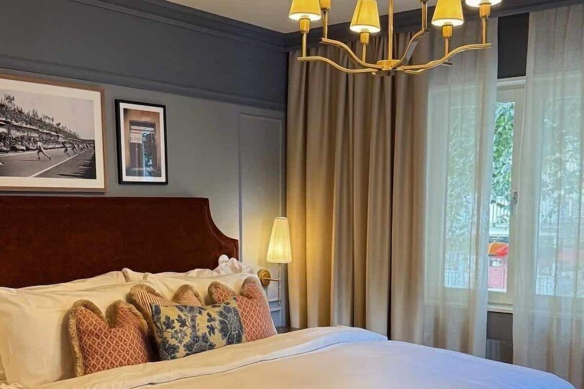 Una habitación de hotel con una cama grande, cabecera de madera, varios almohadones, lámpara de noche, candelabro y una ventana con cortinas.