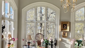 Un comedor con una mesa preparada para dos personas, rodeada de sillas, una araña y ventanas amplias con detalles decorativos.