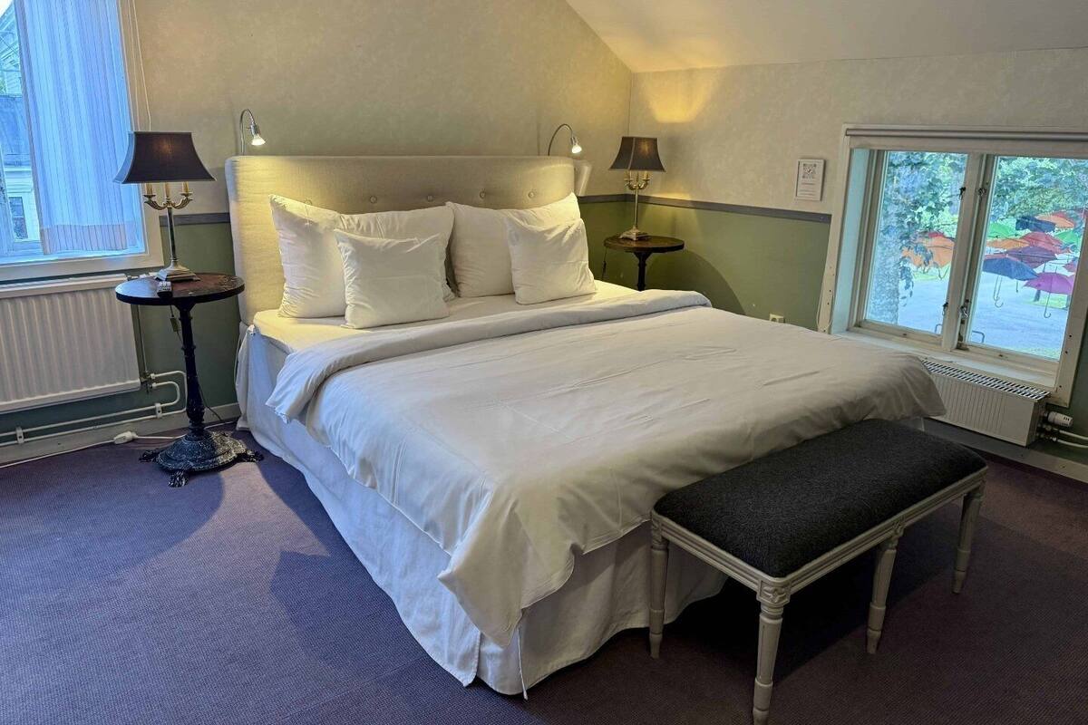 Un dormitorio ordenado con una cama grande, lámparas de noche, un banco y vistas al exterior a través de una ventana.