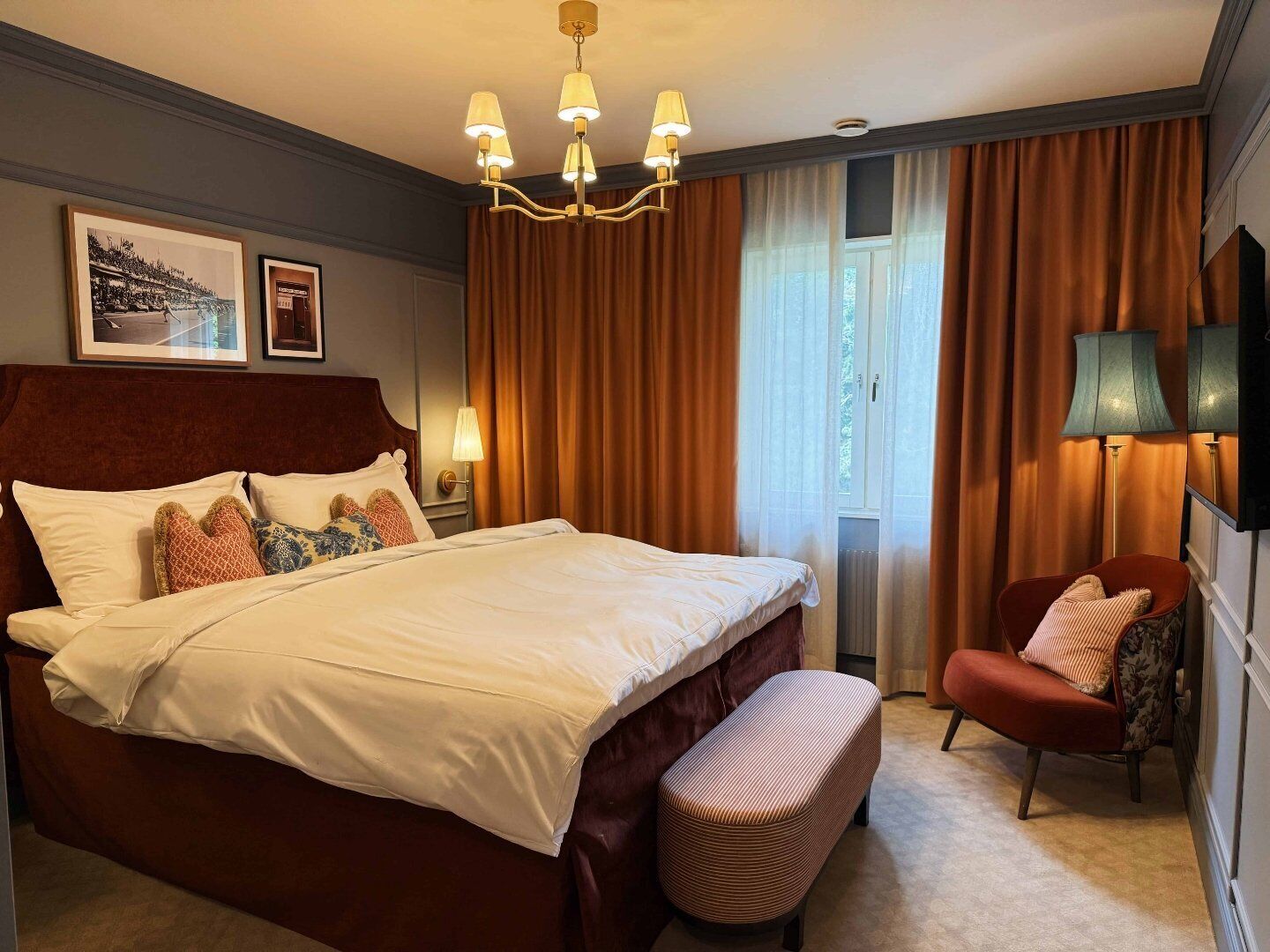Een hotelkamer met een groot bed, een bankje, een stoel, een kroonluchter en gordijnen.
