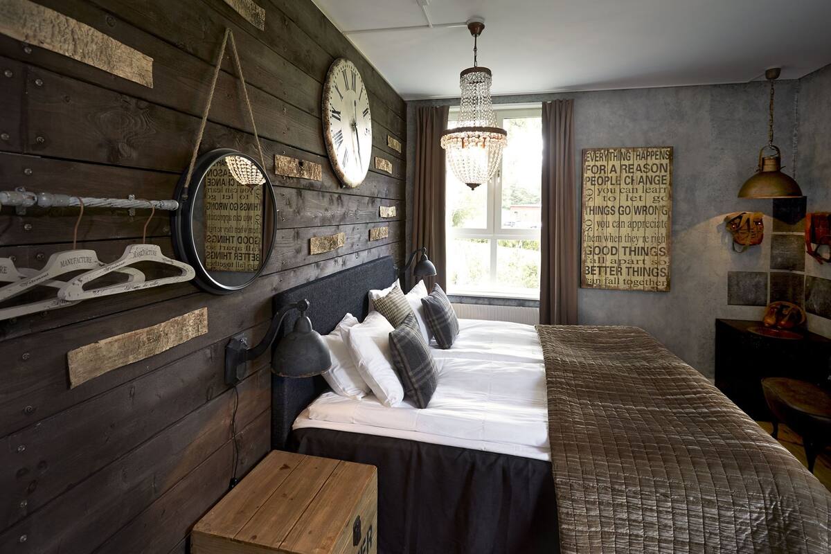 Un dormitorio rústico con pared de madera, una cama con muchos cojines, una lámpara colgante y un reloj de pared grande.