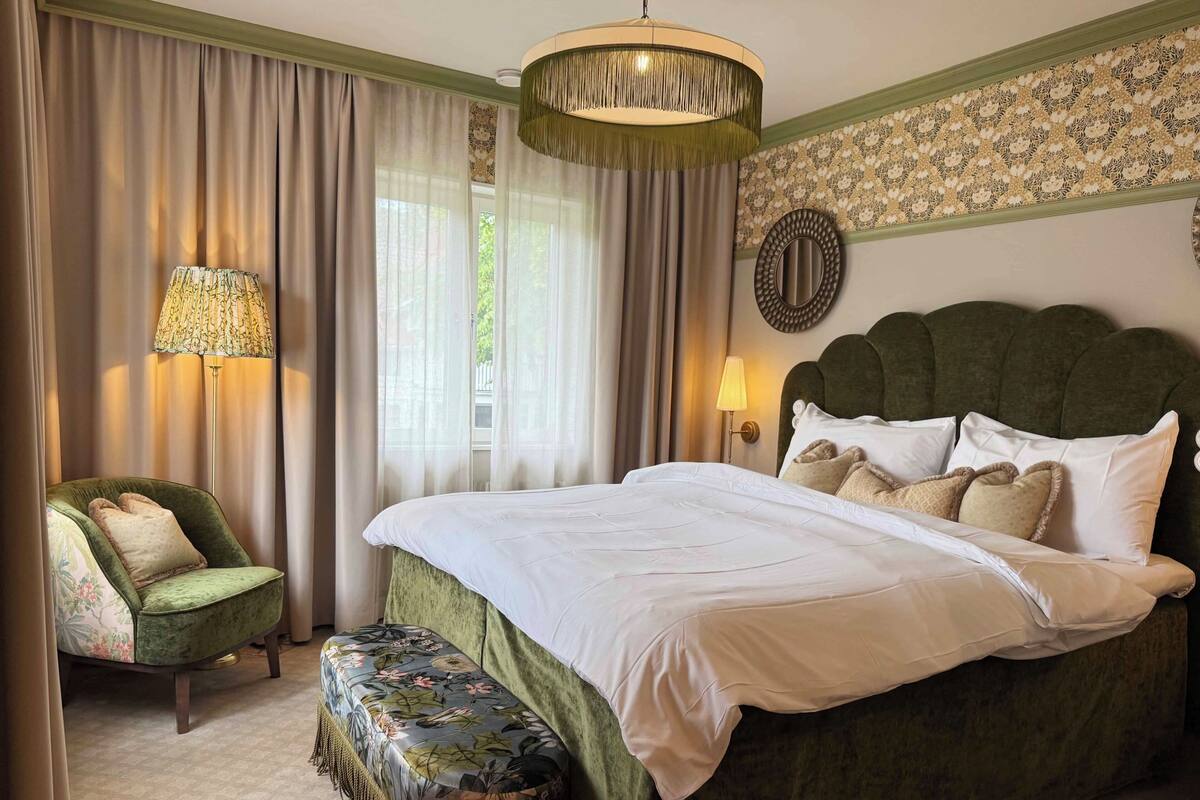 Un dormitorio con una cama grande, un sillón tapizado en verde, un banco con tapicería floral, una lámpara de pie y un espejo montado en la pared.