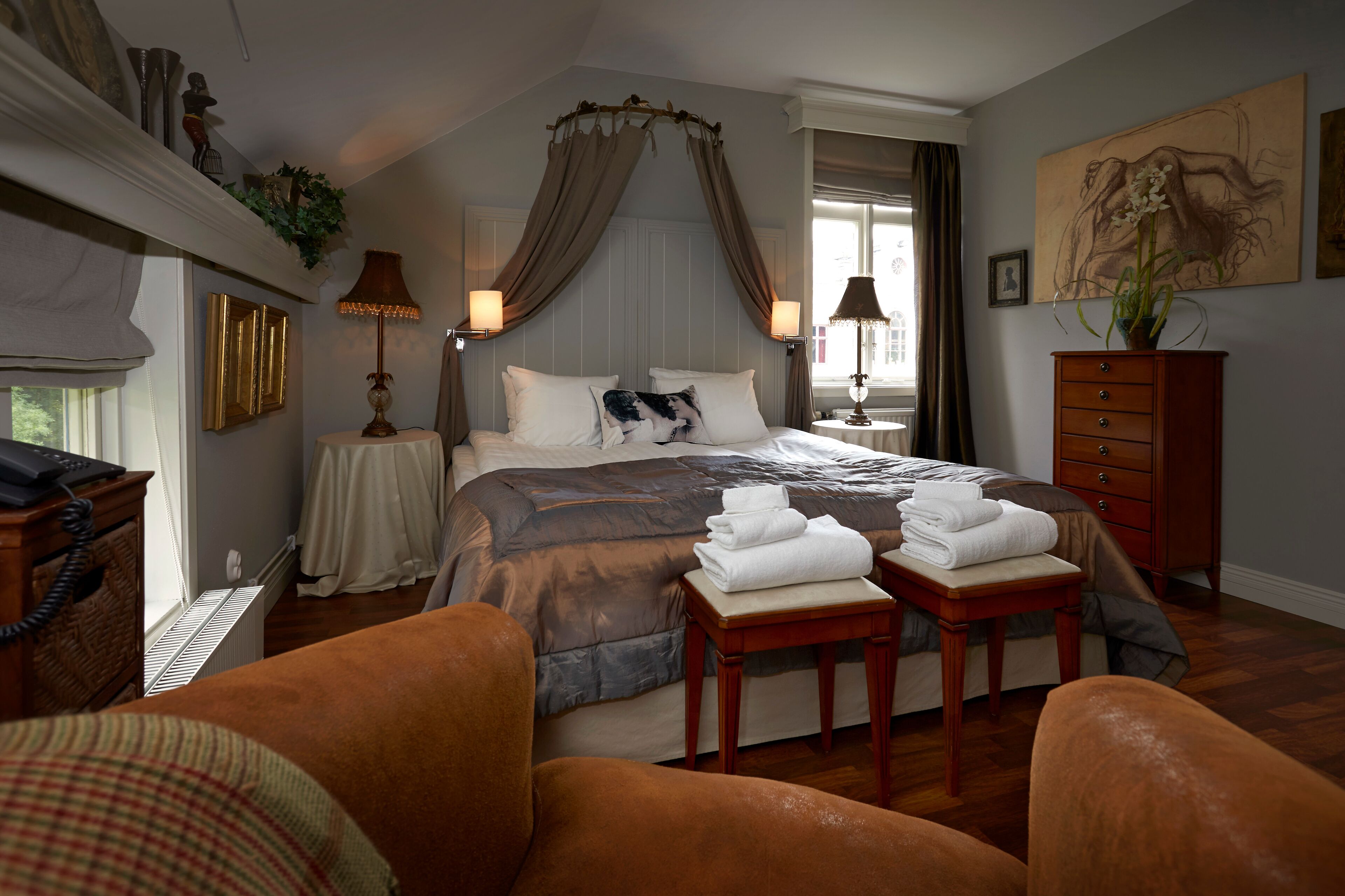 A bedroom with a large bed, a canopy, a nightstand with a lamp, a dresser, and a painting on the wall.