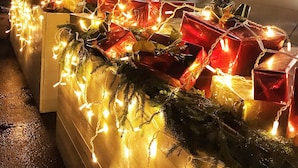Una fila de regalos envueltos con luces, colocados frente a un edificio con un amplio ventanal y una guirnalda en la pared.