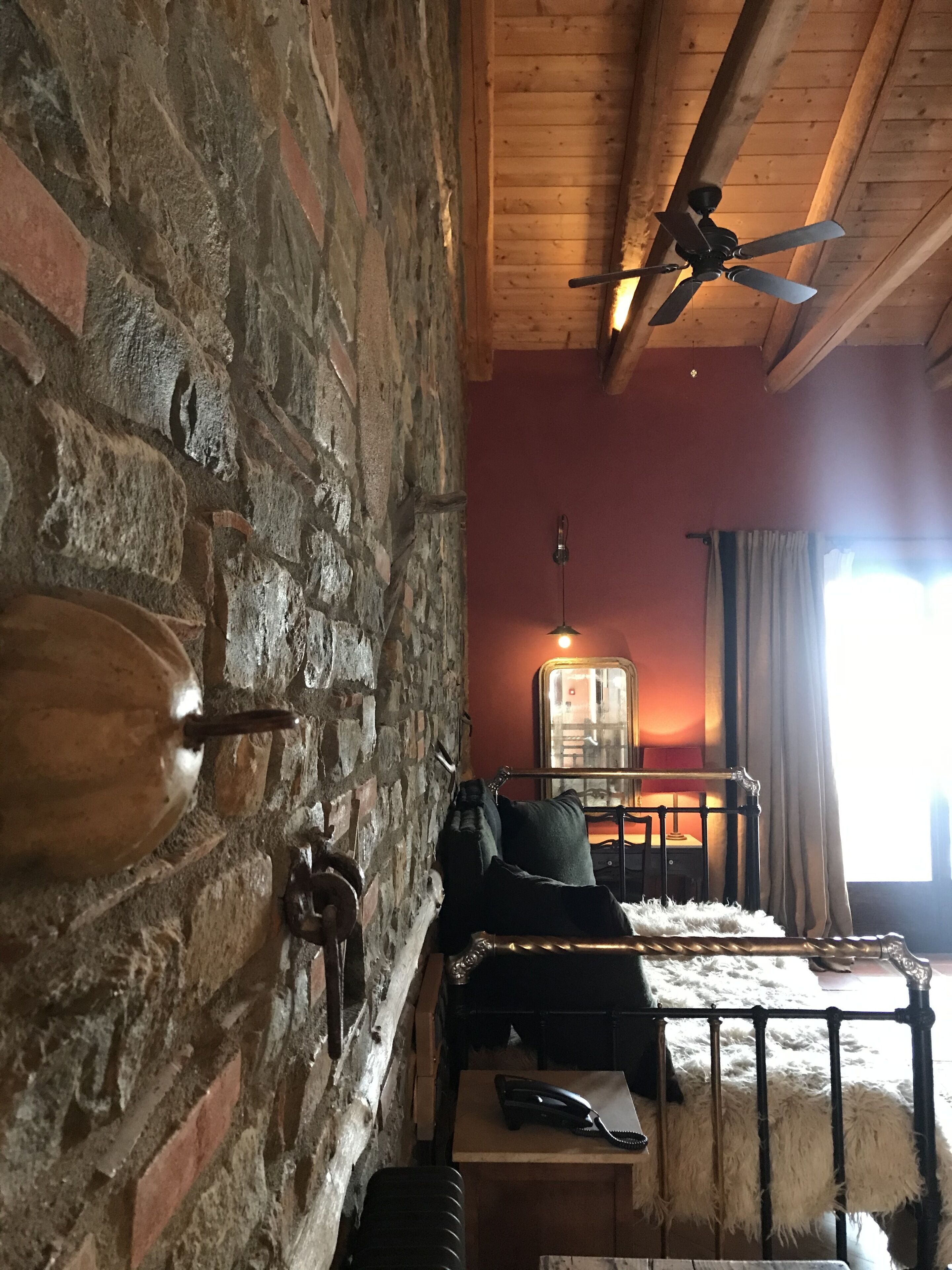 A rustic room with exposed brick walls, a wooden ceiling, a bed with a fluffy white cover, a bedside table with a lamp, and a window with curtains.