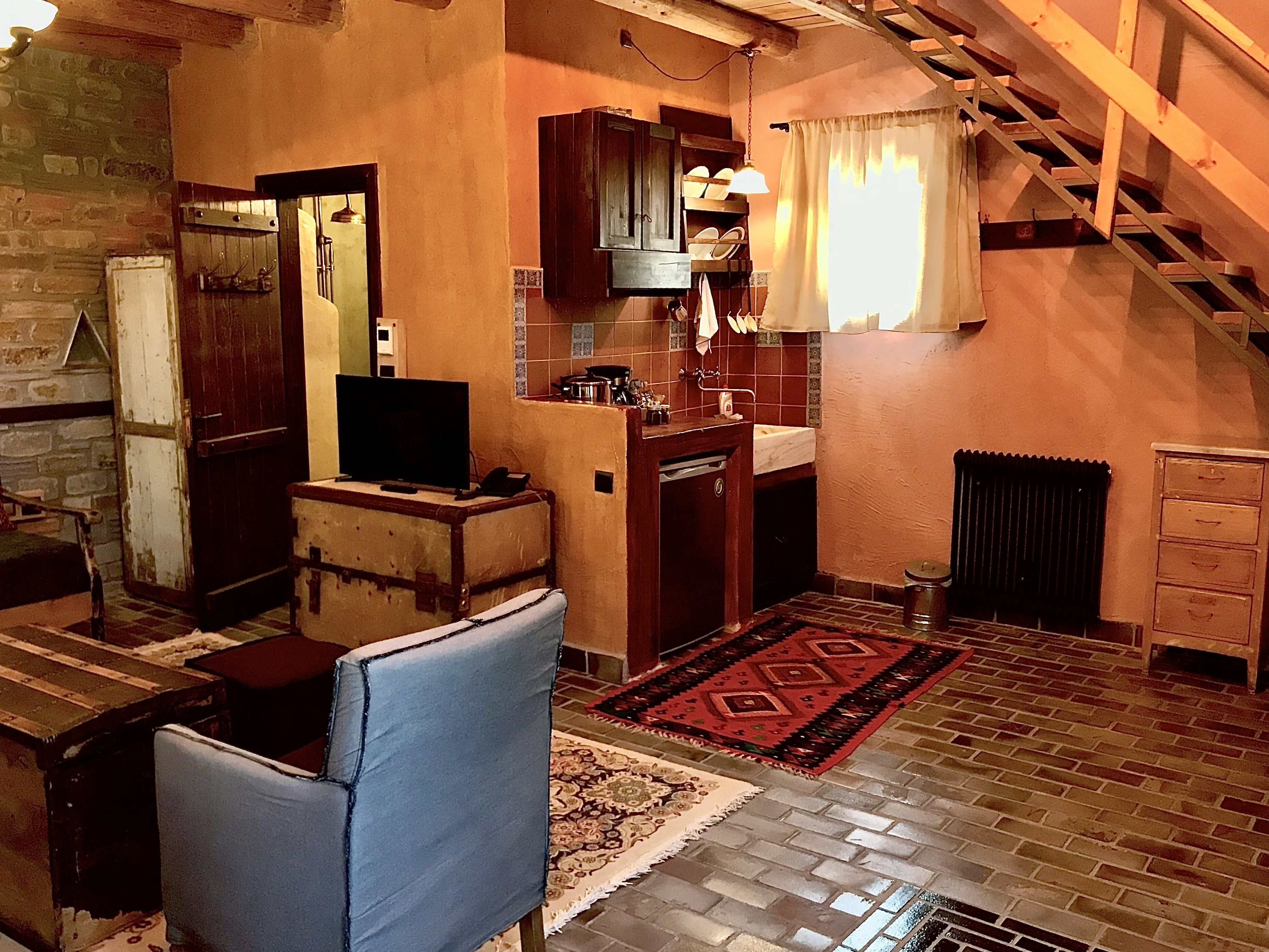 A cozy living room with a television, a blue chair, a kitchen area with cabinets and a sink, and a staircase leading to an upper level.