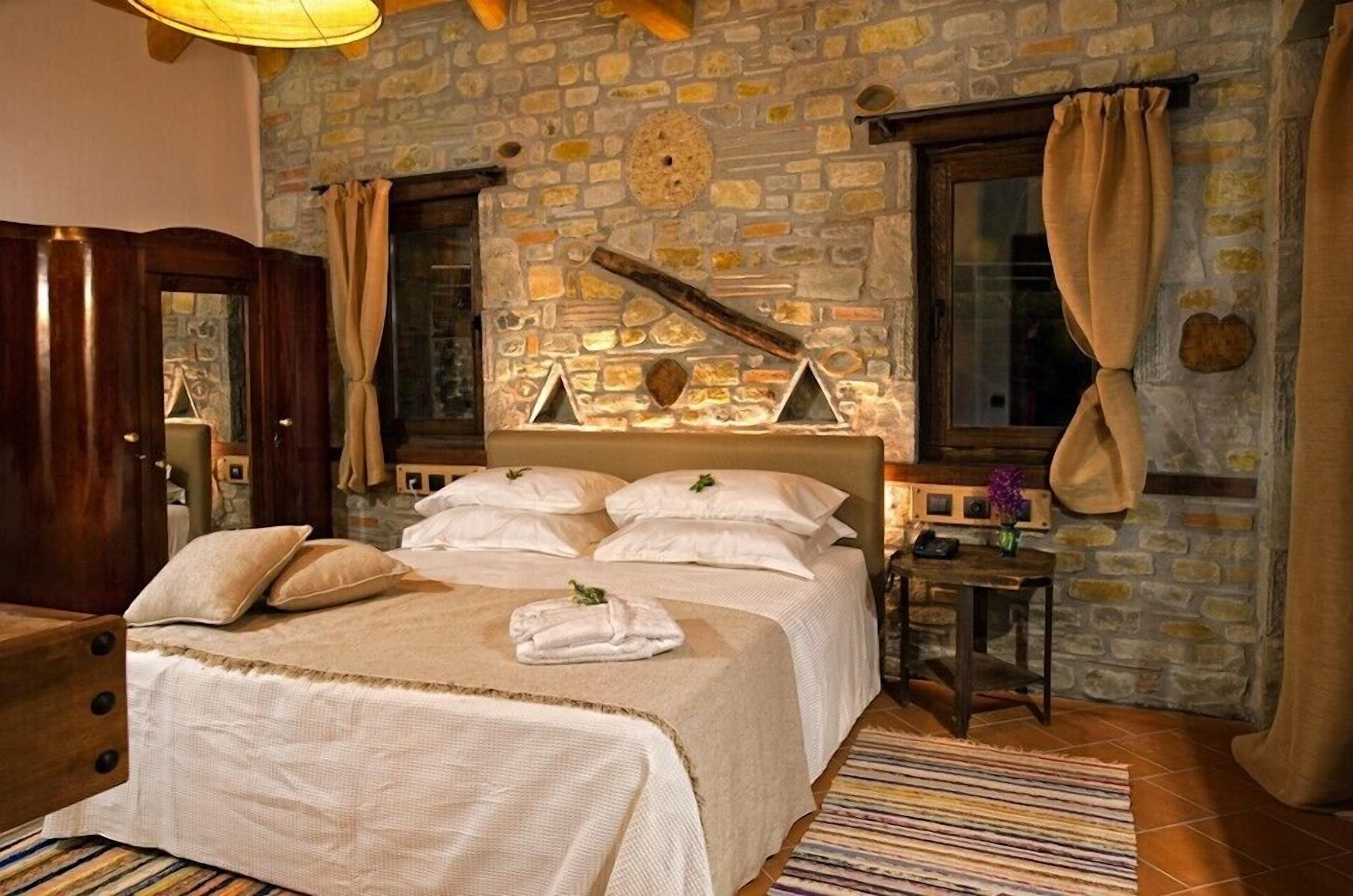 A bedroom with a stone wall, a bed with white linens, a wooden headboard, a bedside table with a lamp, and a window with curtains.