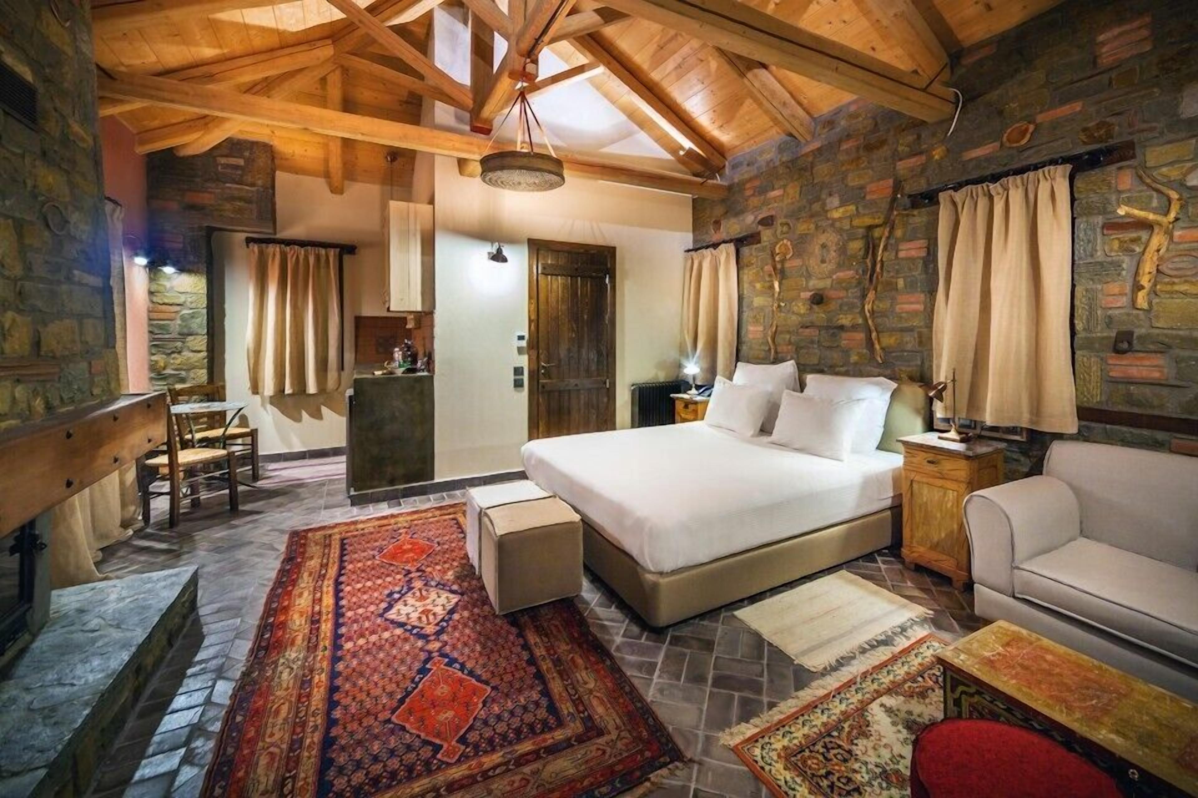A cozy bedroom with a large bed, a wooden bench, a small table, and a red chair. There is a stone wall and a wooden ceiling.