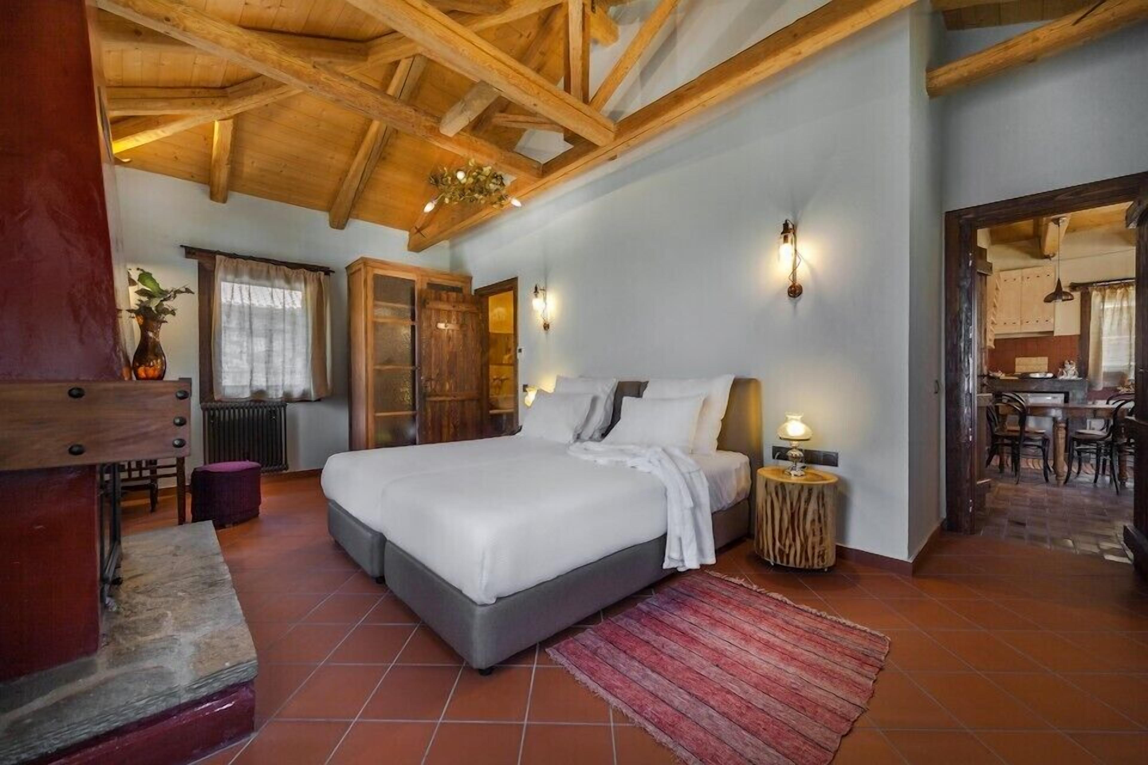 Ein Schlafzimmer mit einem großen Bett, Holzbalkendecke, einem kleinen Tisch mit Lampe und Blick in den Küchenbereich.