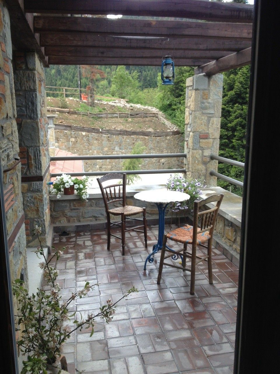 Un patio con paredes de piedra, una mesa redonda y dos sillas, un farol, y vista a una zona boscosa.
