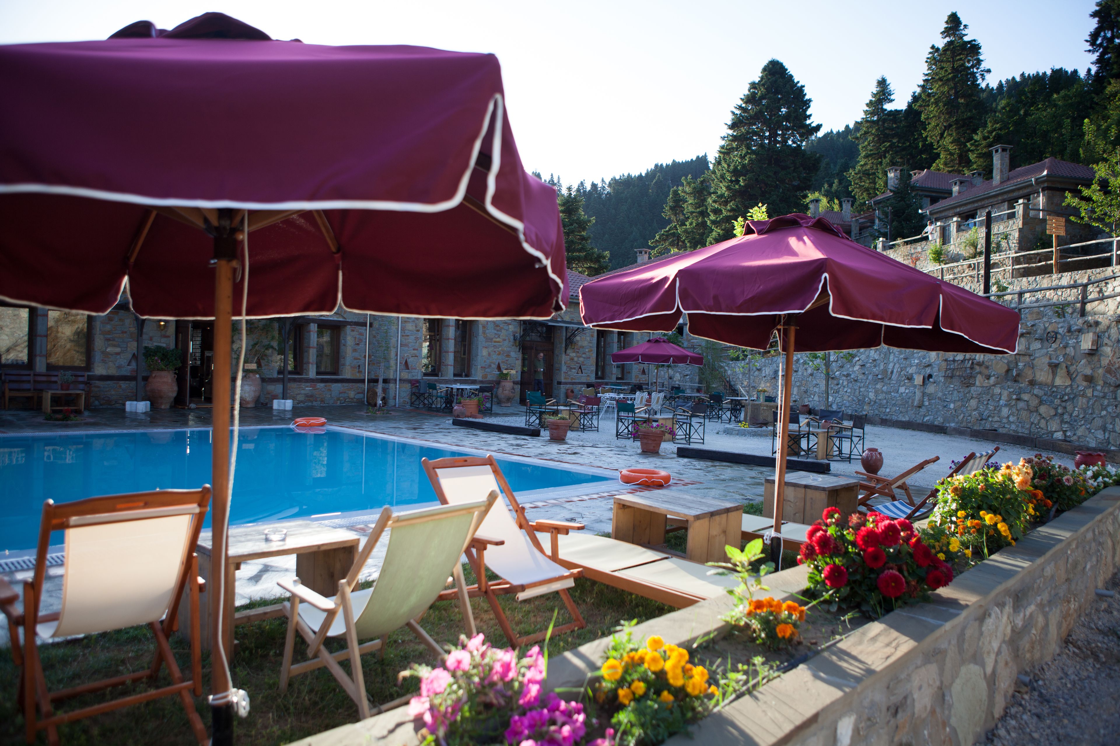 Un área junto a la piscina con sillones reclinables y sombrillas, rodeada de muros de piedra y macizos de flores.