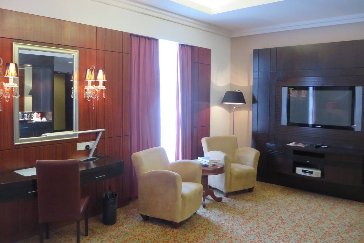 A hotel room with a desk, two armchairs, a TV, a mirror, and a lamp.