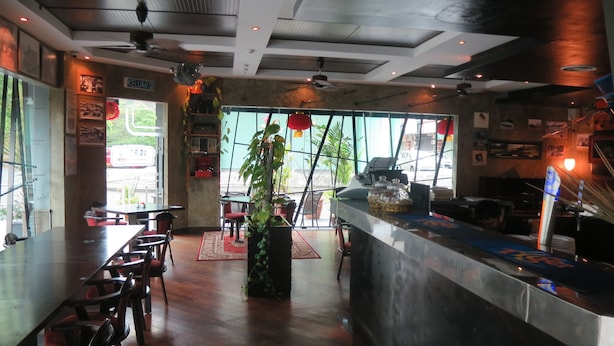 A restaurant interior with wooden tables, chairs, a central plant, and a counter with a menu display. - The Mark's Lodge (Sandakan)