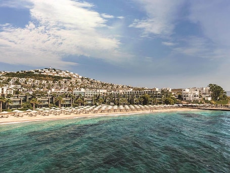 A coastal resort with a beachfront, palm trees, and a hillside with buildings.