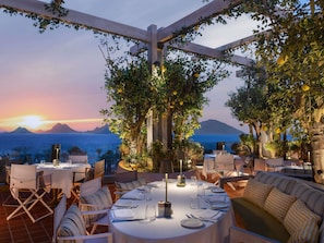 An outdoor dining area with tables and chairs, surrounded by greenery and overlooking a scenic view of the sea and mountains at sunset. - Swissotel Resort Bodrum Beach (Bodrum)