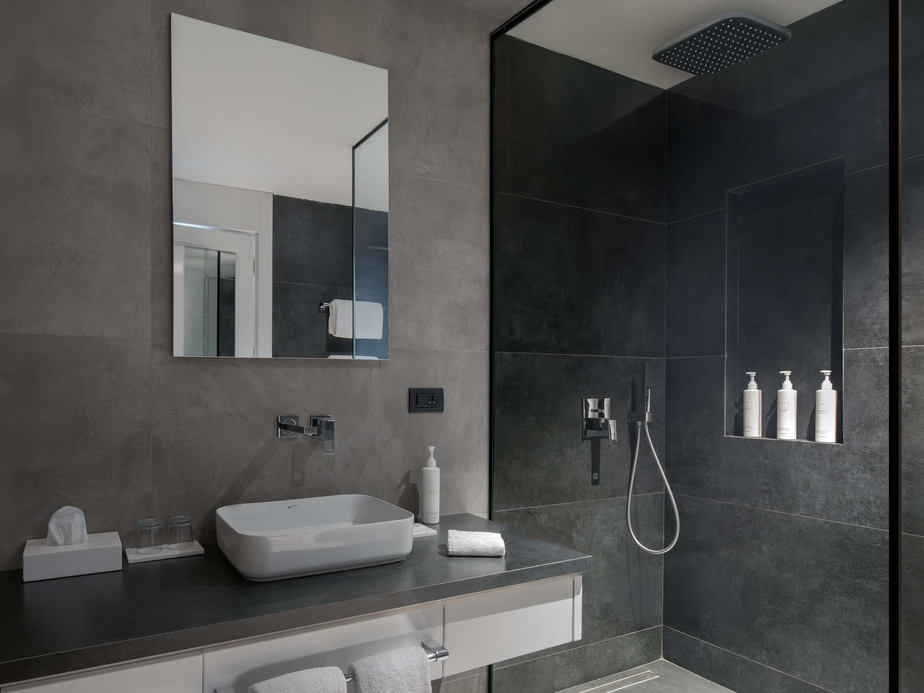 Un baño moderno con un espejo grande, un lavamanos rectangular, un cabezal de ducha de mano y un dispensador de jabón de pared.