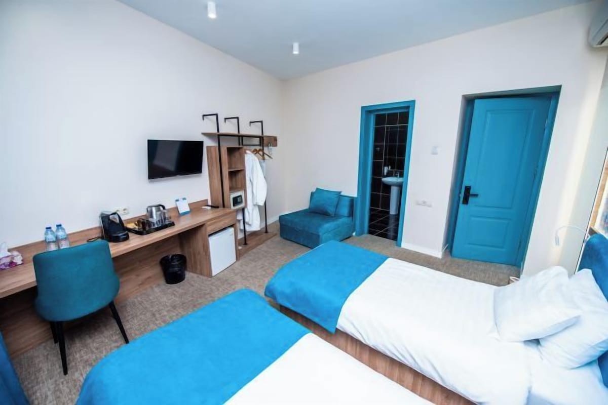 A hotel room with two single beds, a desk with a chair, a TV, and a small kitchenette.