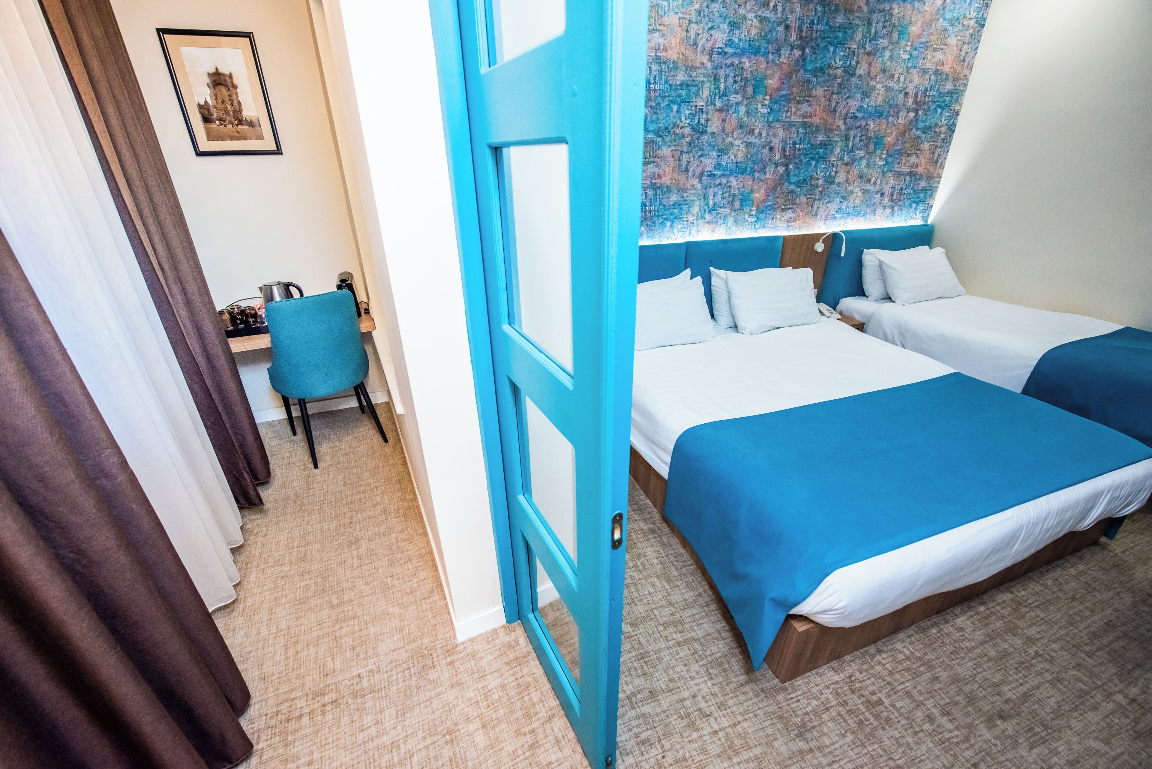 Ein Hotelzimmer mit zwei Betten, einem Schreibtisch mit Stuhl und einem großen Fenster mit einem blauen, abstrakten Gemälde.