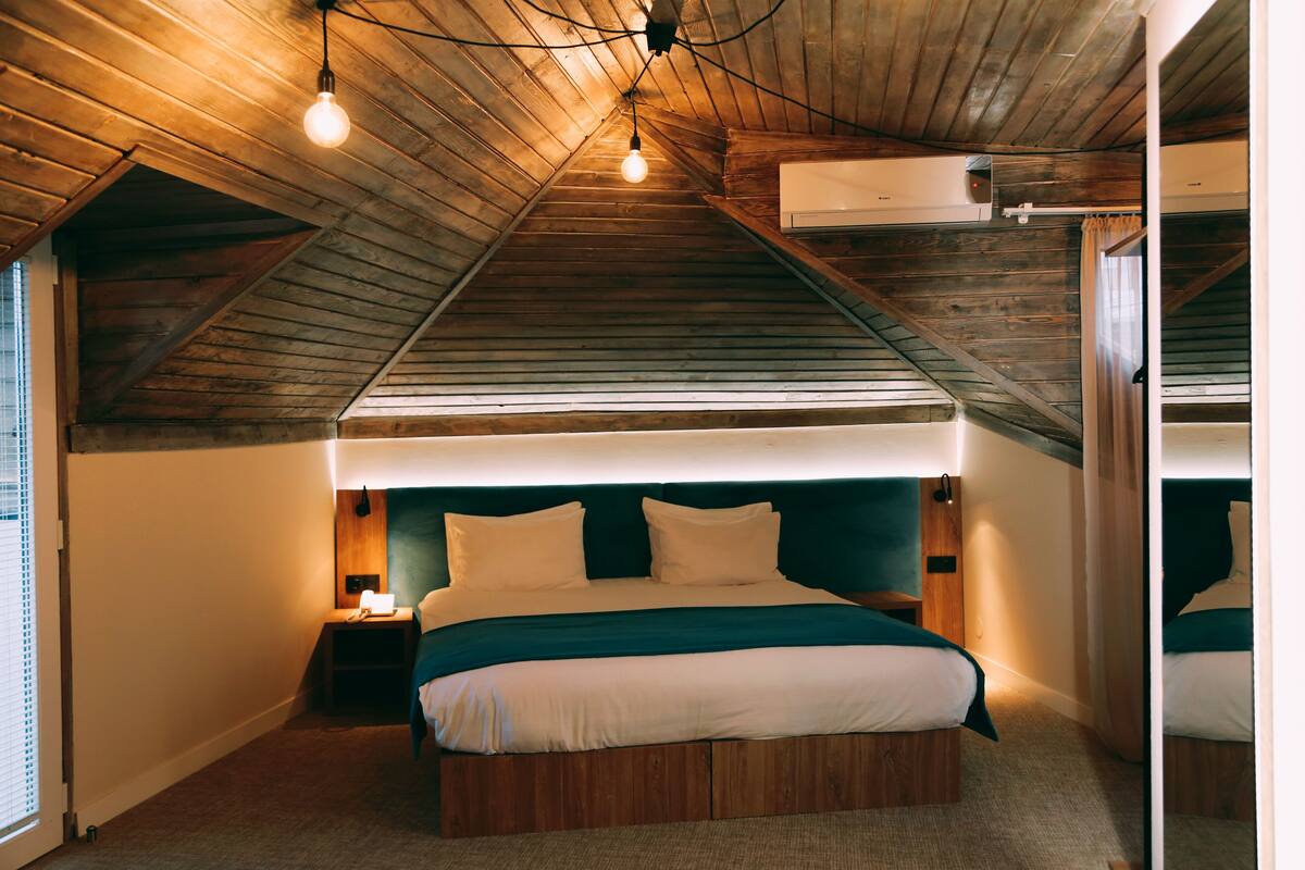 A bedroom with a sloped ceiling, wooden beams, a bed with white bedding, a nightstand, and a wall-mounted air conditioner.