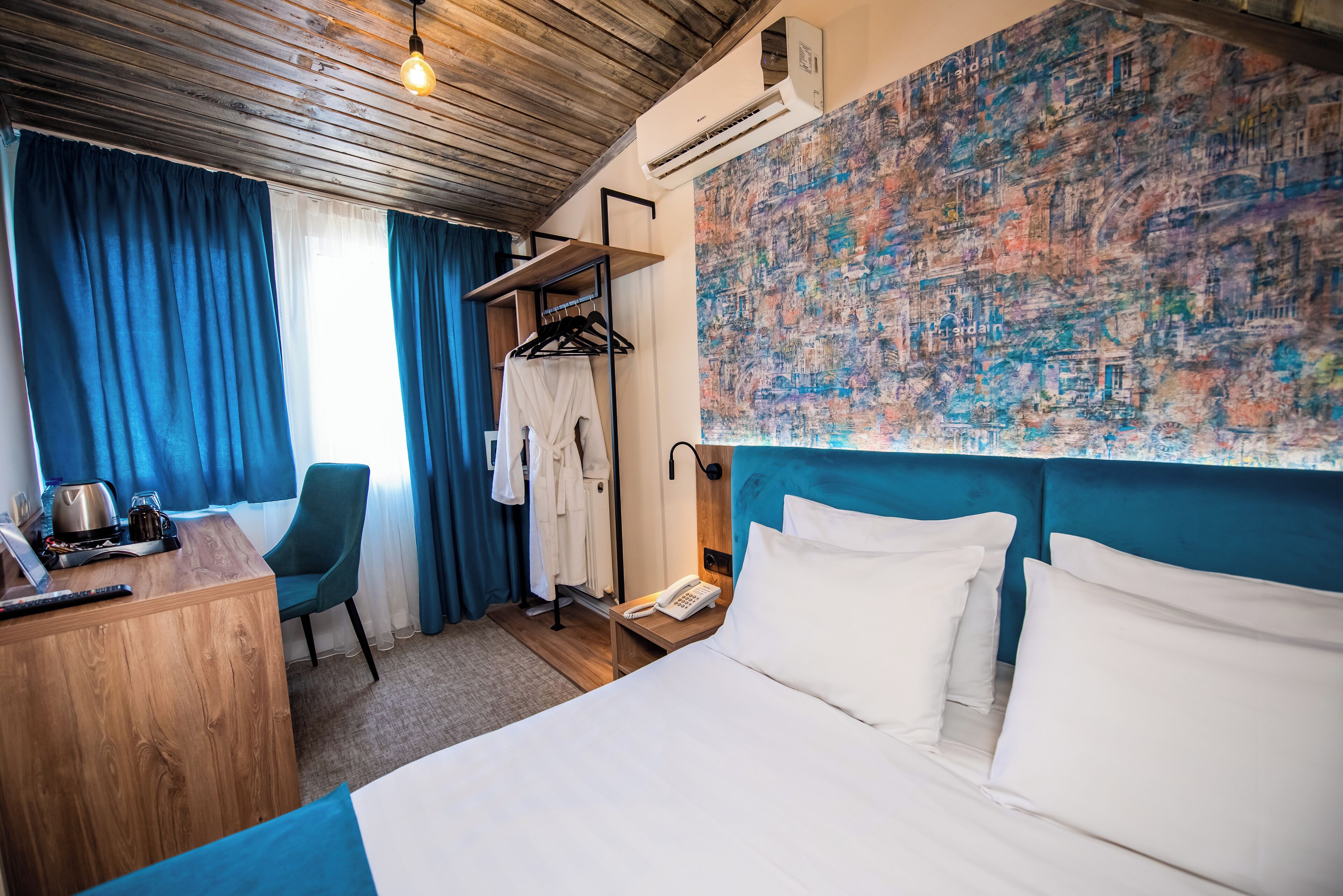 Une chambre d’hôtel avec un bureau en bois, une chaise, un lit avec du linge de lit blanc, une tête de lit bleue, un climatiseur fixé au mur et un grand tableau abstrait.