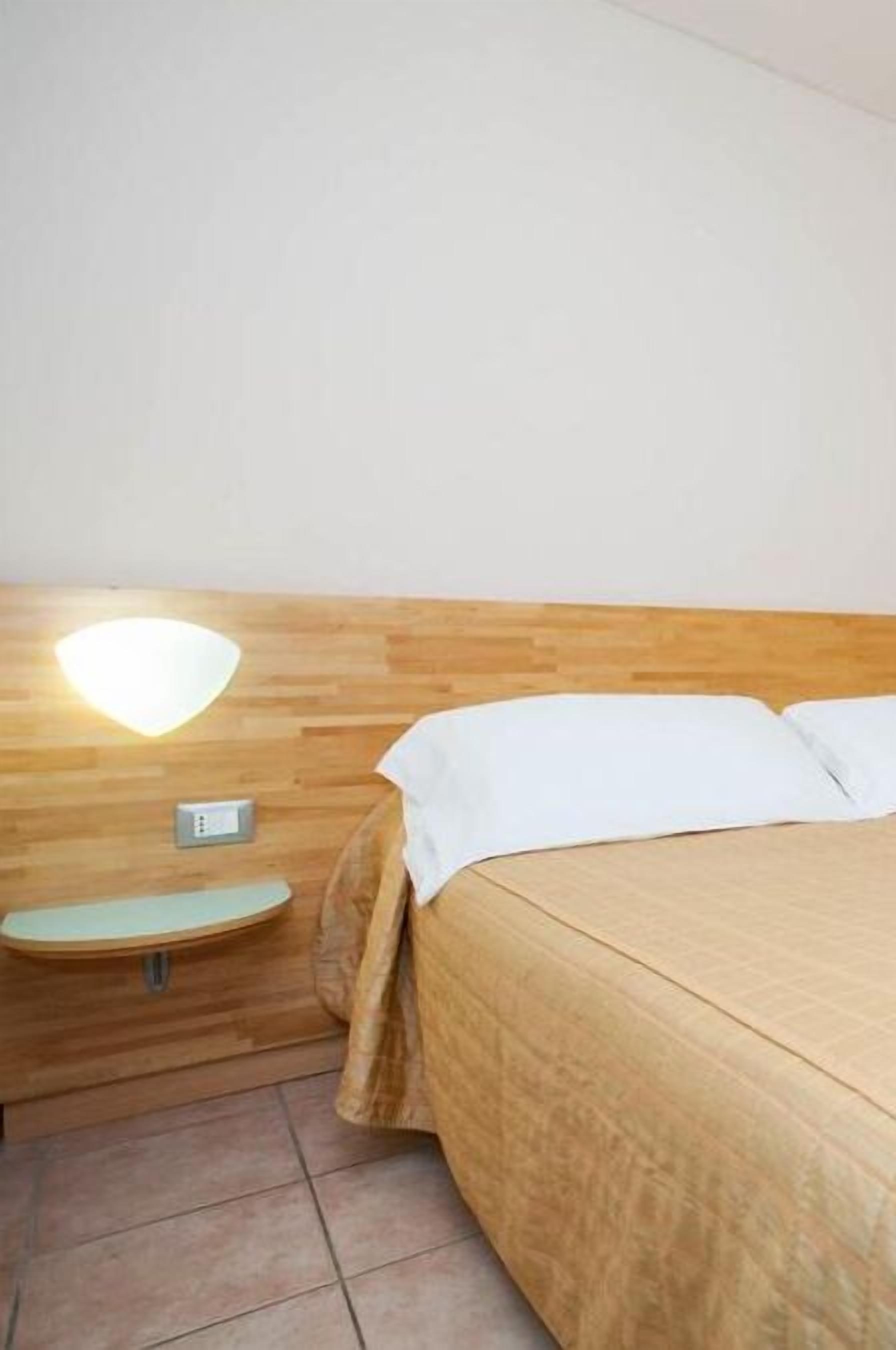Camera d'albergo con testiera in legno, un comodino con una lampada e un letto con lenzuola bianche.