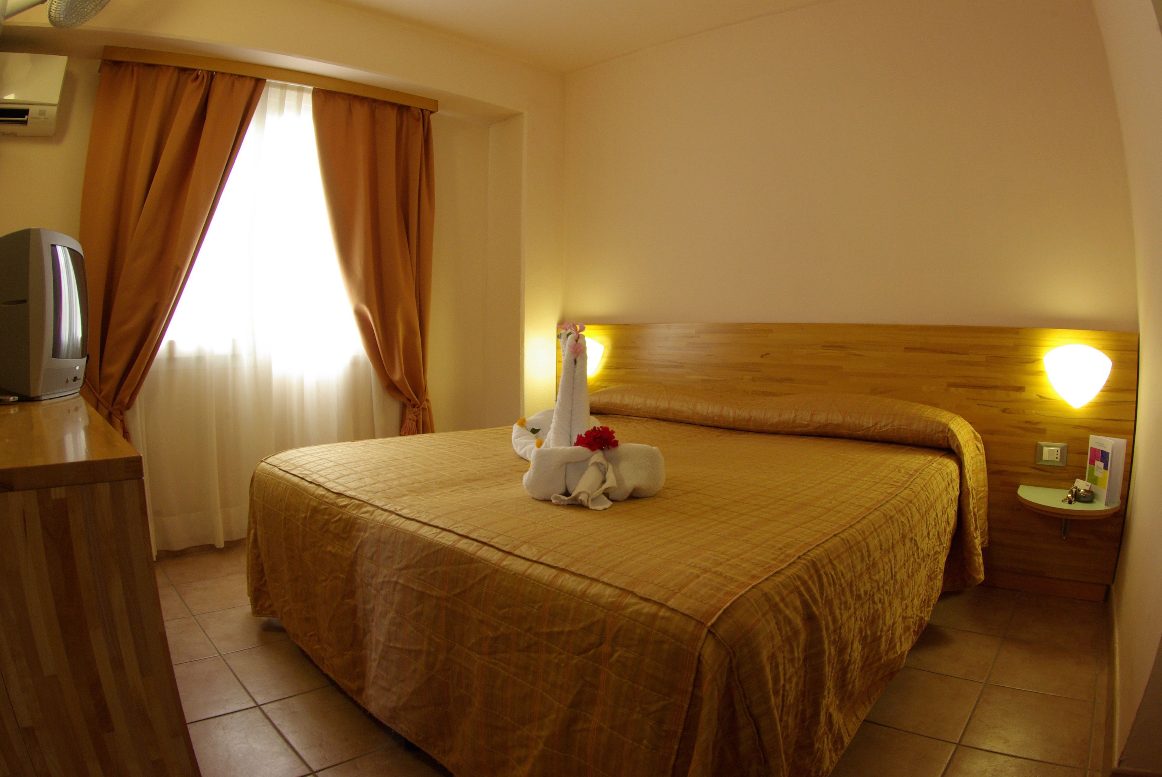 Una camera d'albergo con un letto, una testiera in legno, un comodino con una lampada, una televisione e tende.