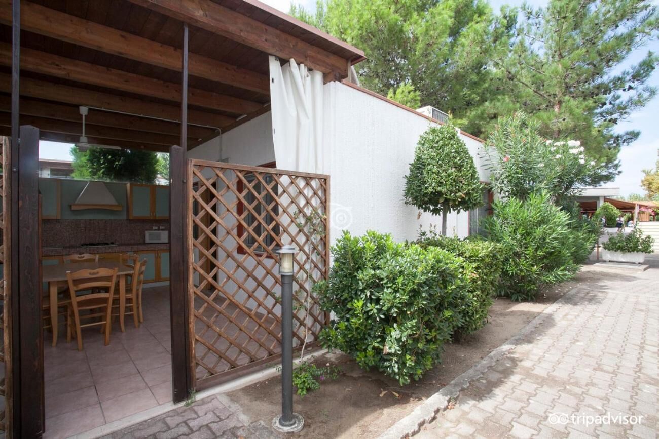 Un'area salotto esterna coperta con tavolo e sedie in legno, una recinzione a griglia e un muro bianco con un piccolo albero.