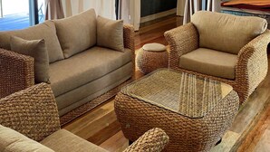 Amplia sala de estar con un conjunto de sofás, una mesa de centro y sillas de mimbre. Un candelabro cuelga del techo, y cortinas cubren los laterales.