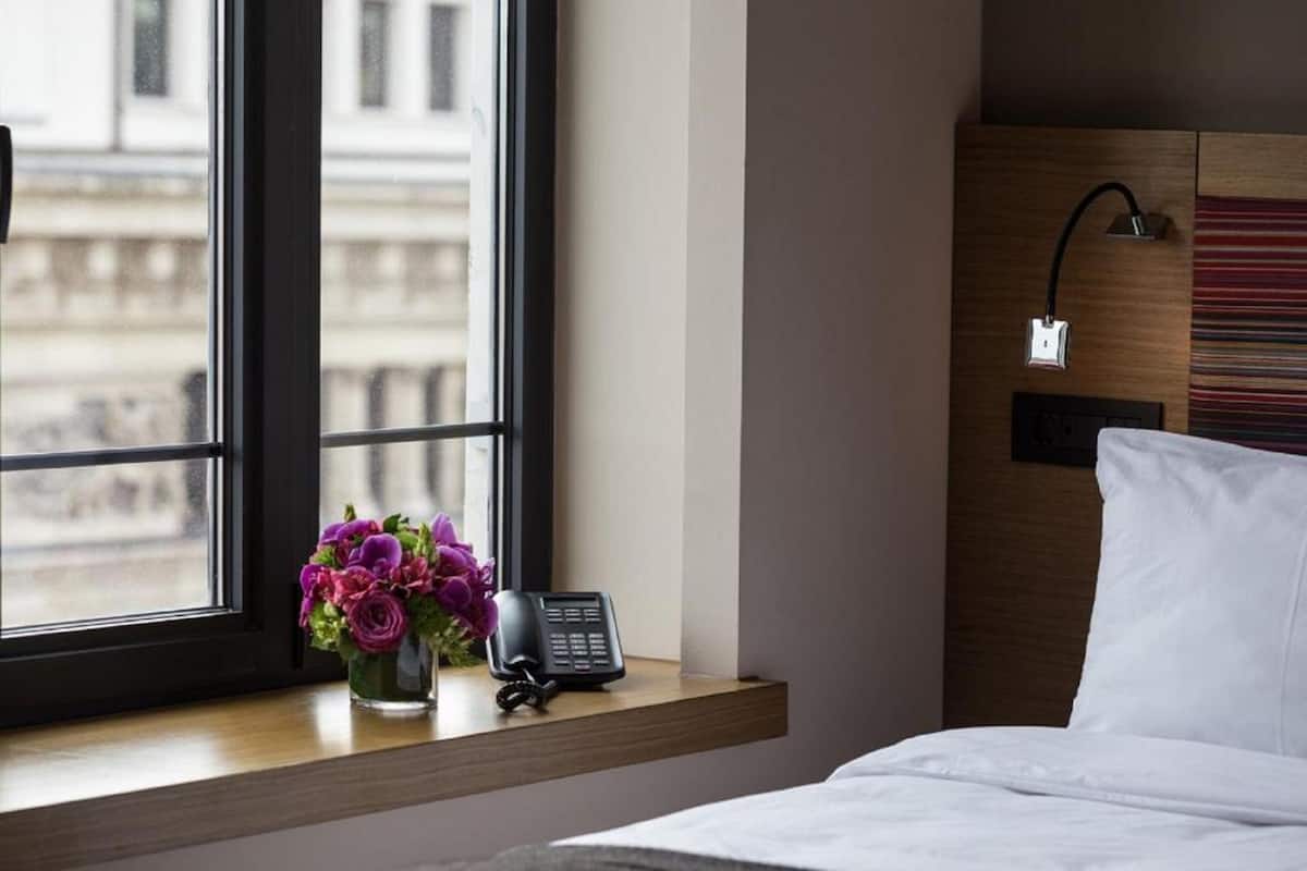 Une chambre d’hôtel avec un lit, une table de chevet avec un téléphone et des fleurs, une fenêtre donnant sur des immeubles et une lampe fixée au mur.