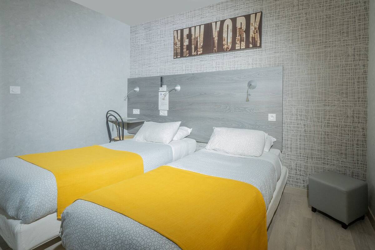 Quarto de hotel com duas camas, cabeceira com arte urbana e uma mesinha de cabeceira, além de um pequeno banquinho.