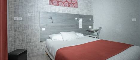 Un dormitorio moderno con cama, una colcha roja, cabecera, lámpara de pared y una mesita.