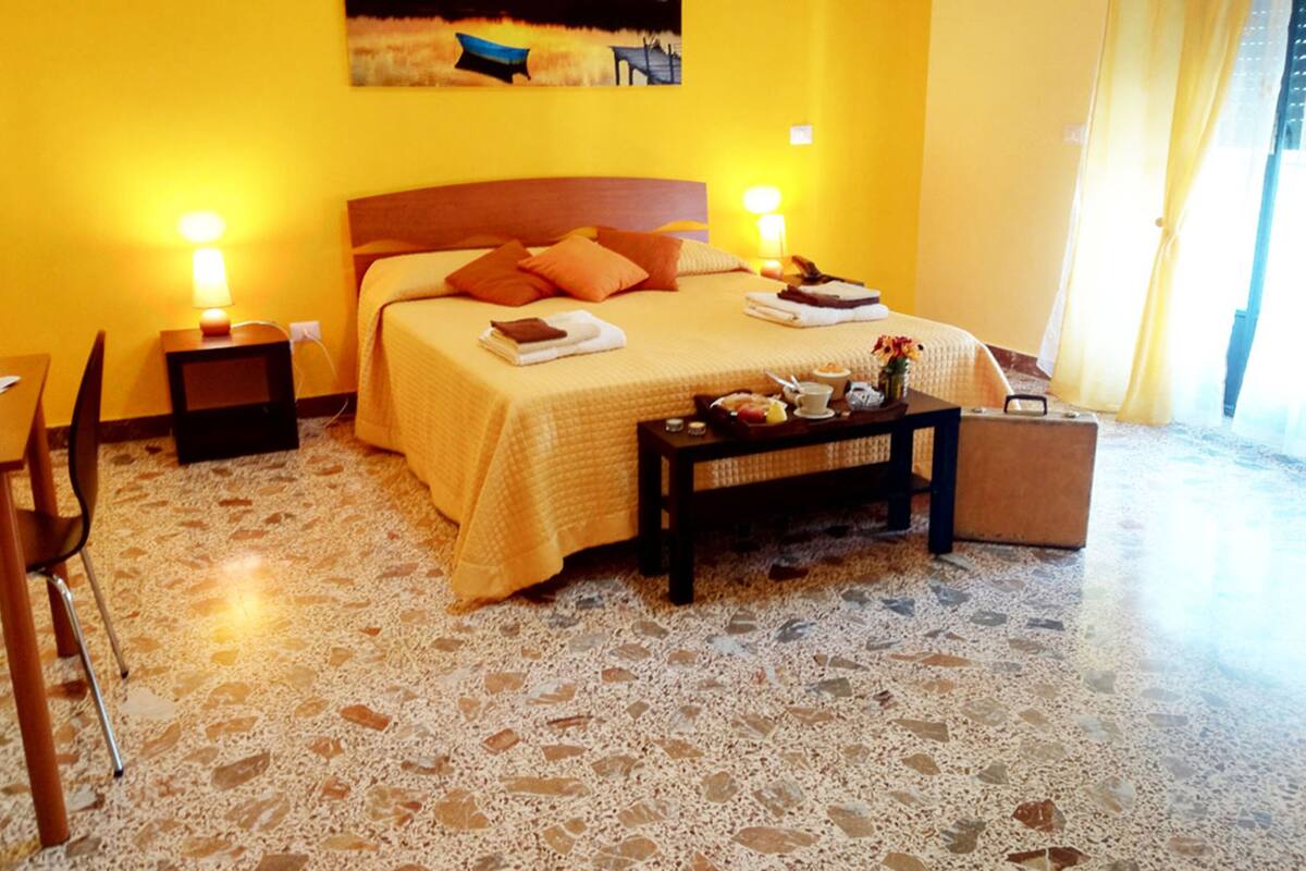 Habitación de hotel con cama, mesitas de noche, una silla y una pequeña mesa con detalles de cortesía.