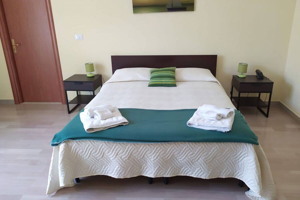 Una cama bien hecha con un almohadón verde, dos mesitas de noche con lámparas y una puerta.