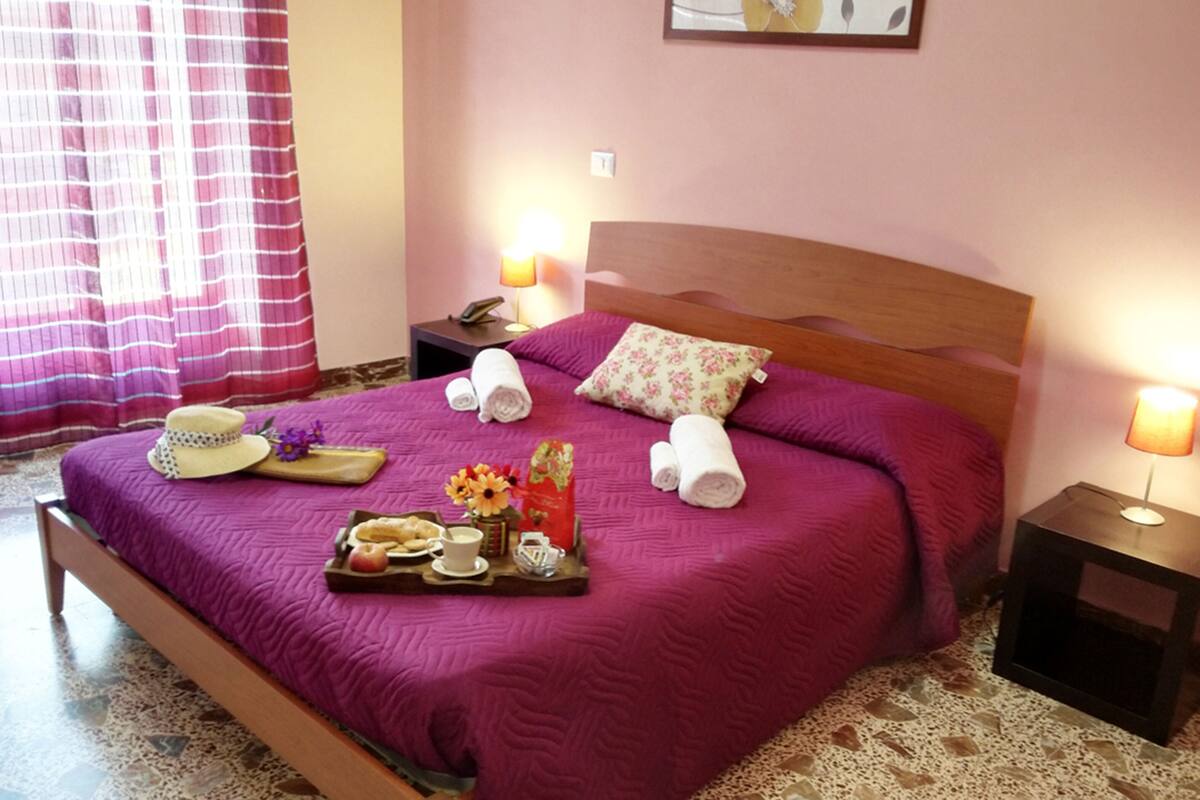 Una cama bien hecha con una colcha morada, dos lámparas de noche, un cabecero de madera y una bandeja con artículos para el desayuno.