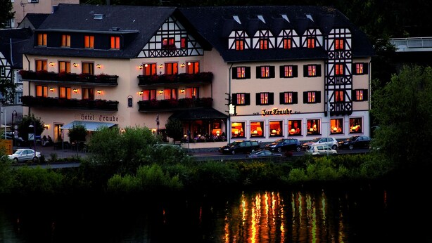 A hotel with a traditional European architectural style, featuring multiple balconies and a river in the foreground. - Hotel Traube (Loef)