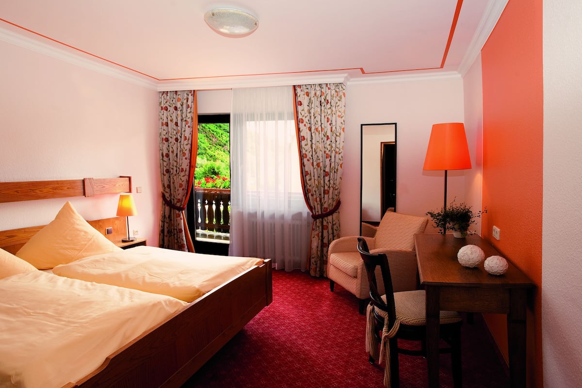 A hotel room with a bed, a desk with a chair, a lamp, and a view of greenery outside.