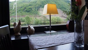 A room with a view of a river and hills, a lamp on a table, and decorative items including a rooster and a vase with a plant. - Hotel Traube (Loef)