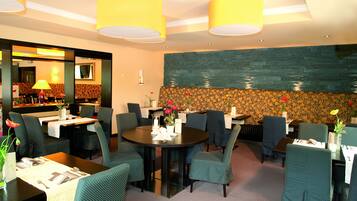 A restaurant with round tables and green upholstered chairs, featuring a stone wall and yellow ceiling lights.