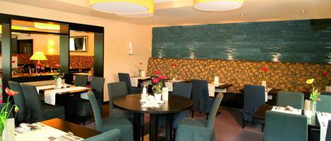 Ein Restaurant mit runden Tischen und grünen Sitzstühlen, das eine Steinwand und gelbe Deckenleuchten hat.