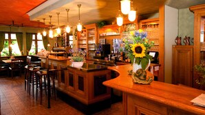 A bar area with wooden furniture, a counter, and a large vase with flowers. - Hotel Traube (Loef)