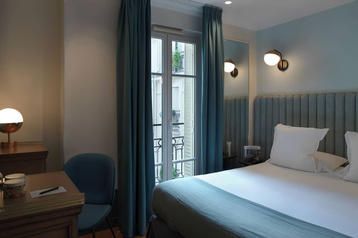 A hotel room with a bed, a desk with a lamp, a chair, and a window with curtains.