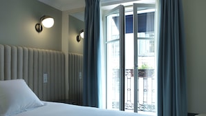 A hotel room with a bed, a wall-mounted light fixture, and a view of a balcony with a railing. - Hotel Bachaumont (Paris)