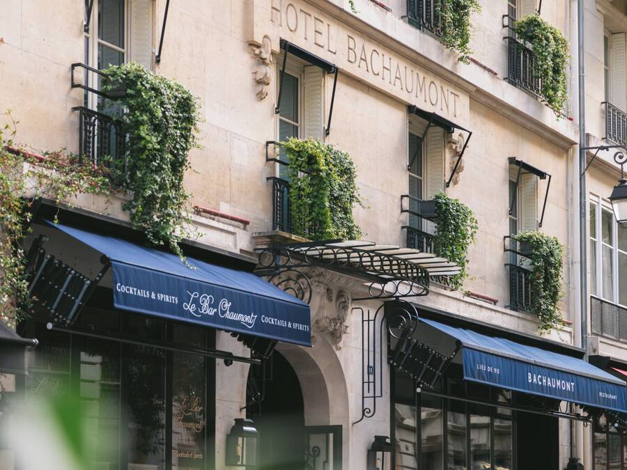 Fachada de hotel con balcones, un letrero que dice 'HOTEL BACHAUMONT' y mobiliario exterior con mesas y sillas.
