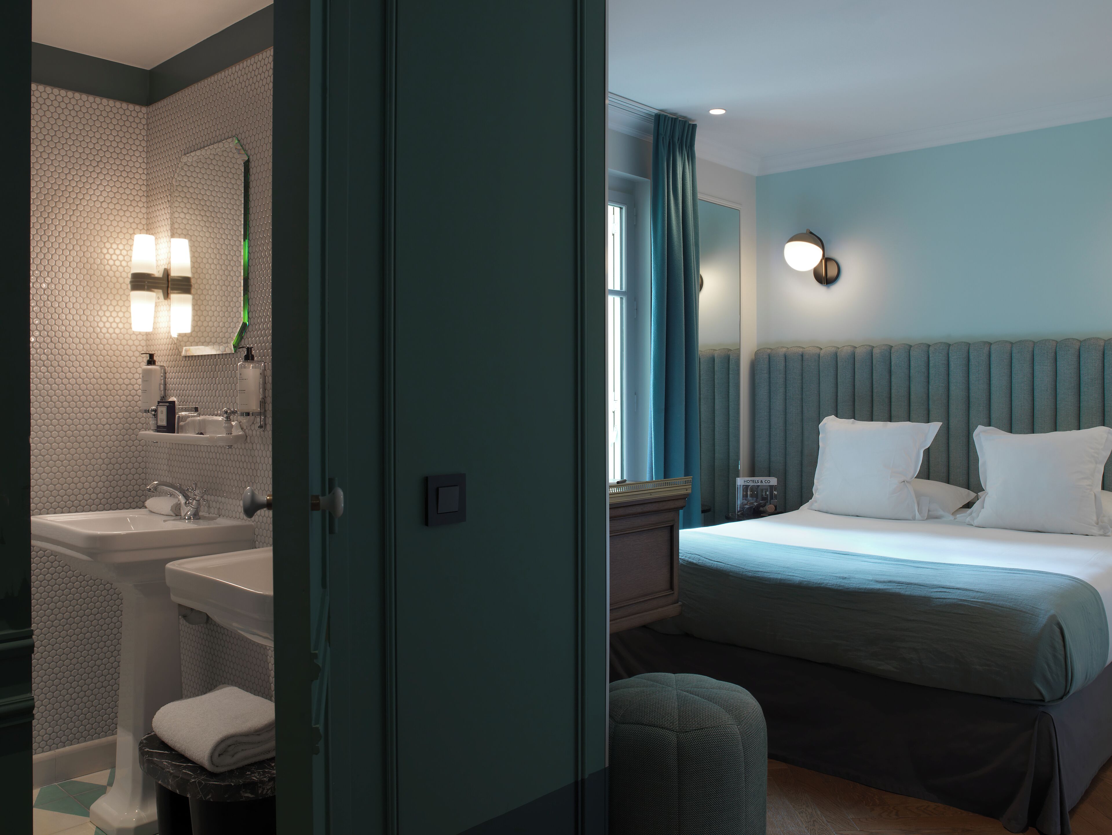 Ett hotellrum med badrum som har handfat, spegel och hygienartiklar. Rummet har en säng med sänggavel, kuddar och ett nattduksbord med en lampa.