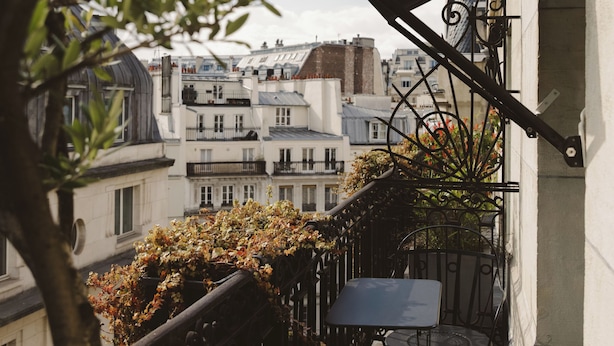 A balcony with a metal railing, a small table, and chairs. There are plants and a tree in the foreground. - Hotel Bachaumont (Paris)