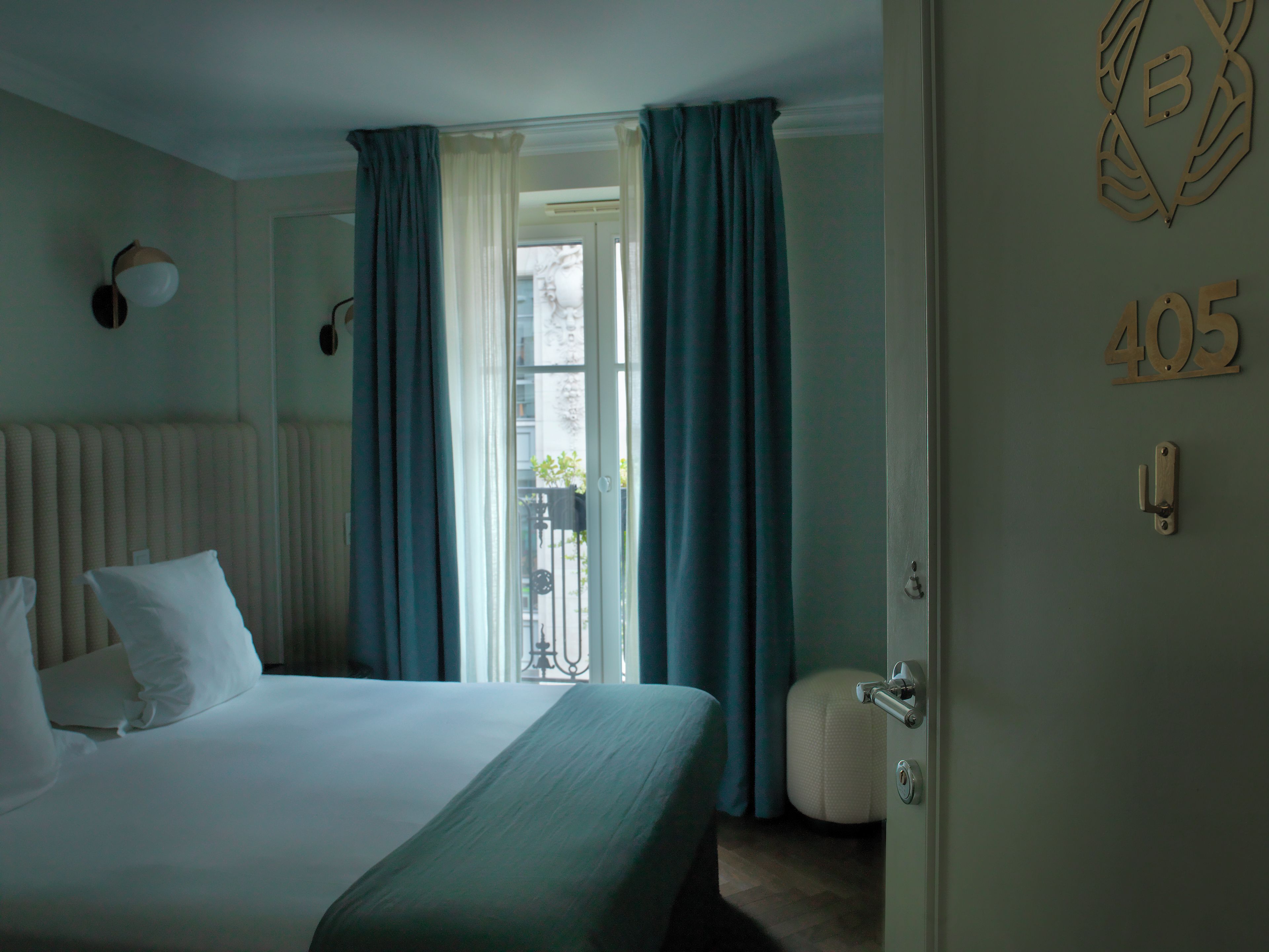 Ein Hotelzimmer mit Bett, Heizkörper, Fenster mit Vorhängen und einer Tür mit einer Nummer.'