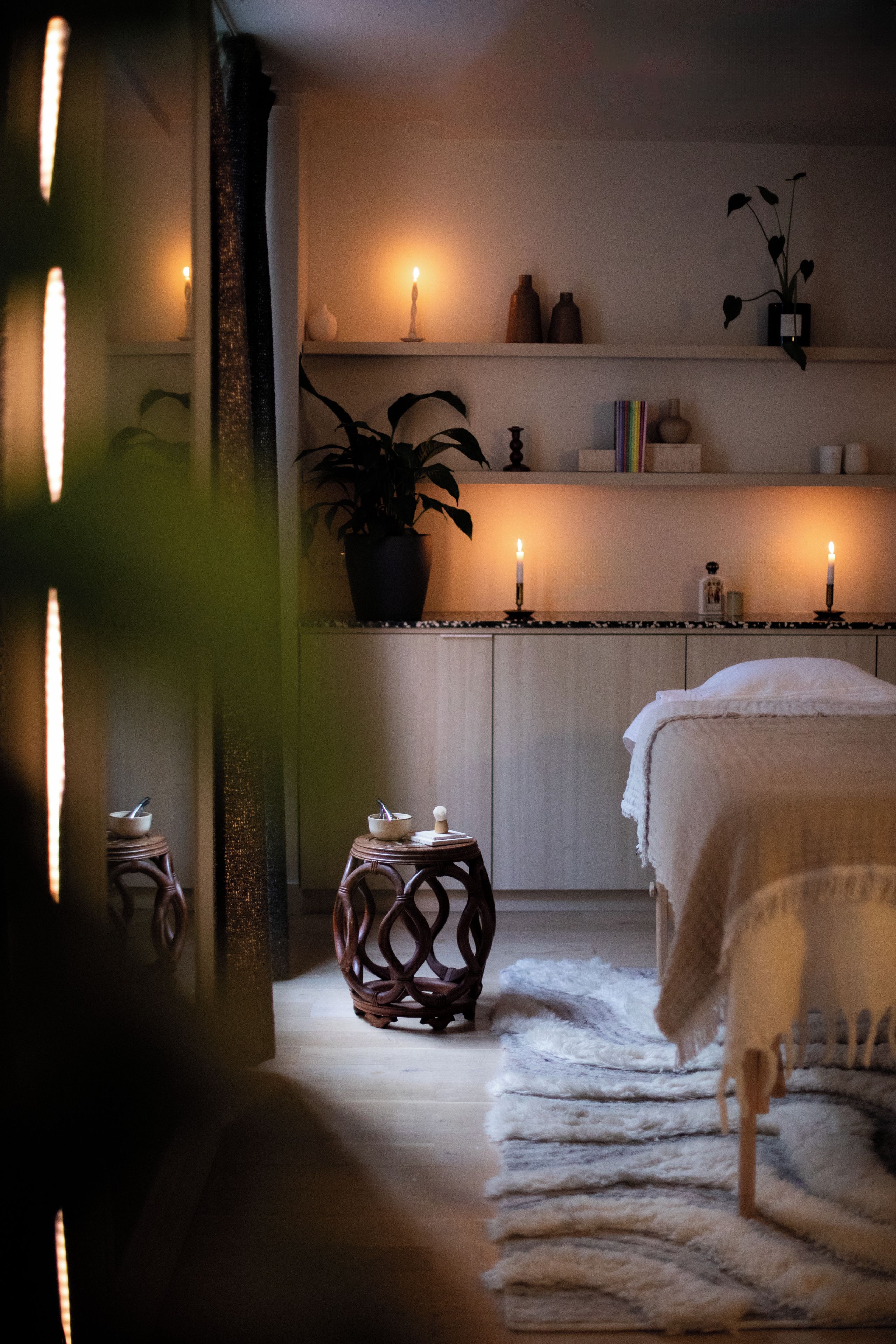 Une pièce confortable avec un lit, une petite table, une plante en pot et une étagère où sont posées des bougies et des livres.