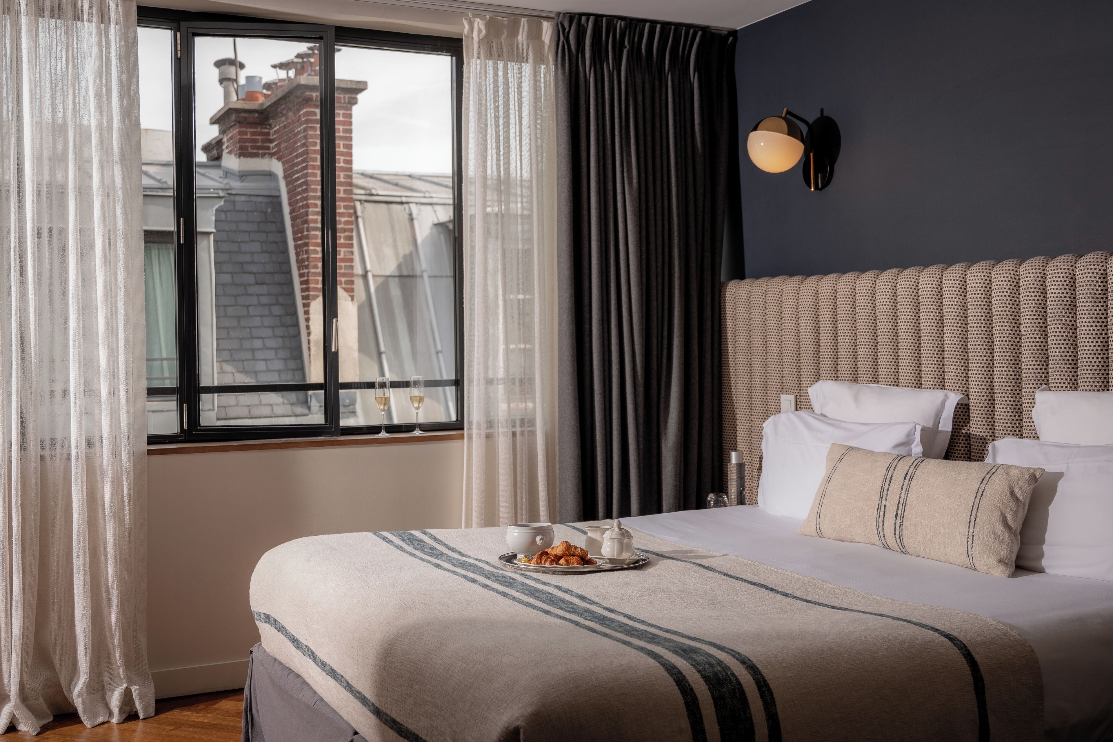 A hotel room with a large bed, a view of the cityscape through the window, and a modern wall-mounted light fixture.